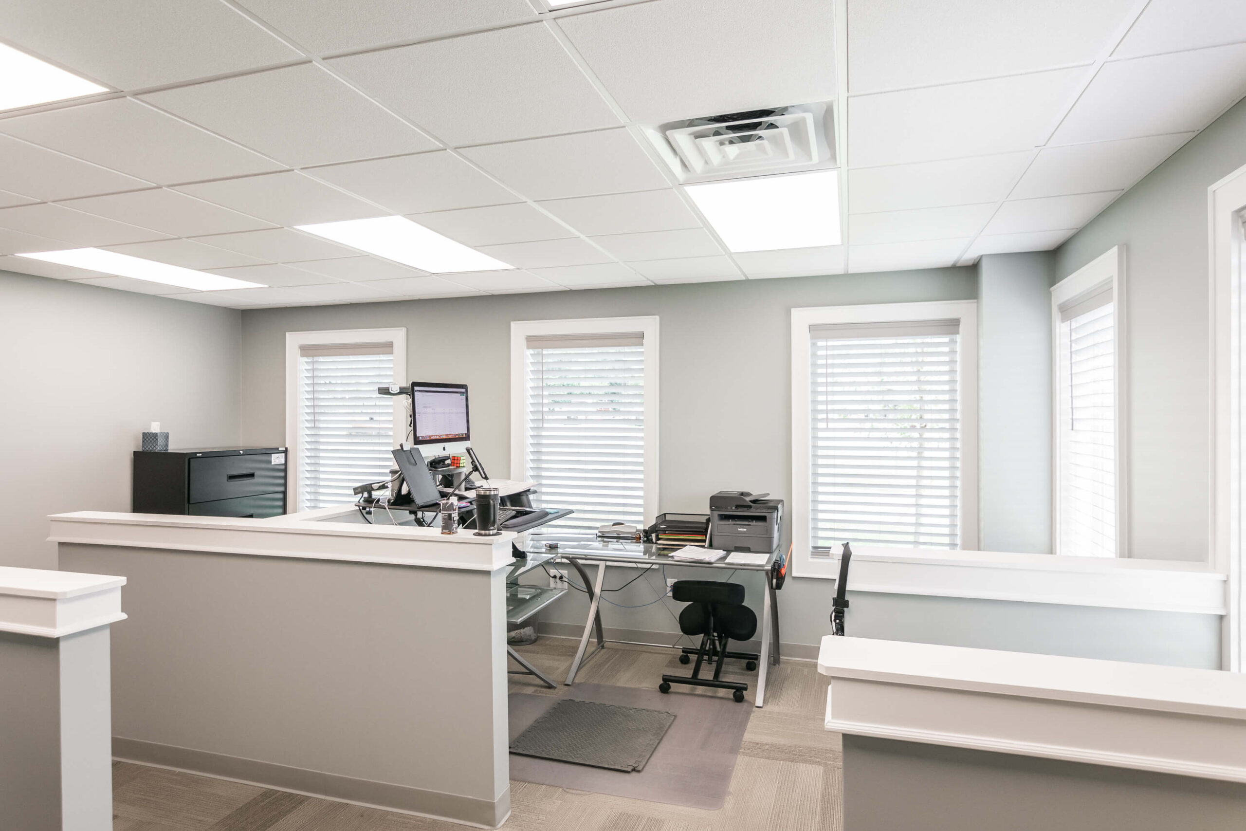 Modern office space with a desk holding a computer, printer, and office supplies. Four windows with blinds line the wall, and overhead lighting brightens the room.