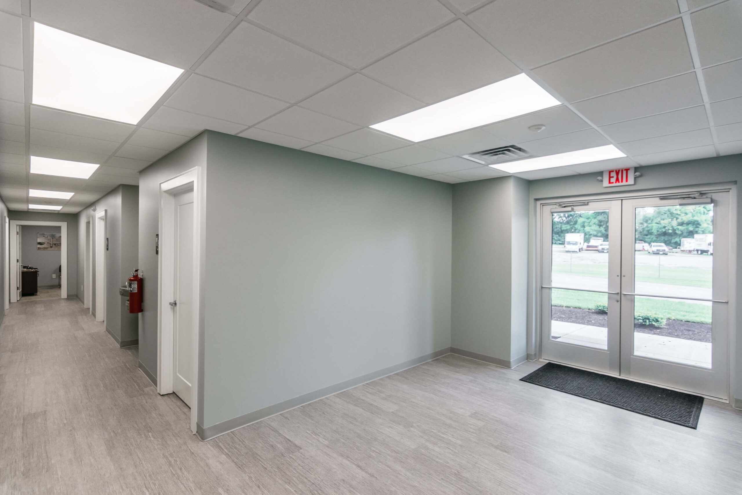 Empty hallway with gray walls, white doors, and a tiled floor. Glass double doors lead outside, with a view of greenery. Fluorescent lights illuminate the space.