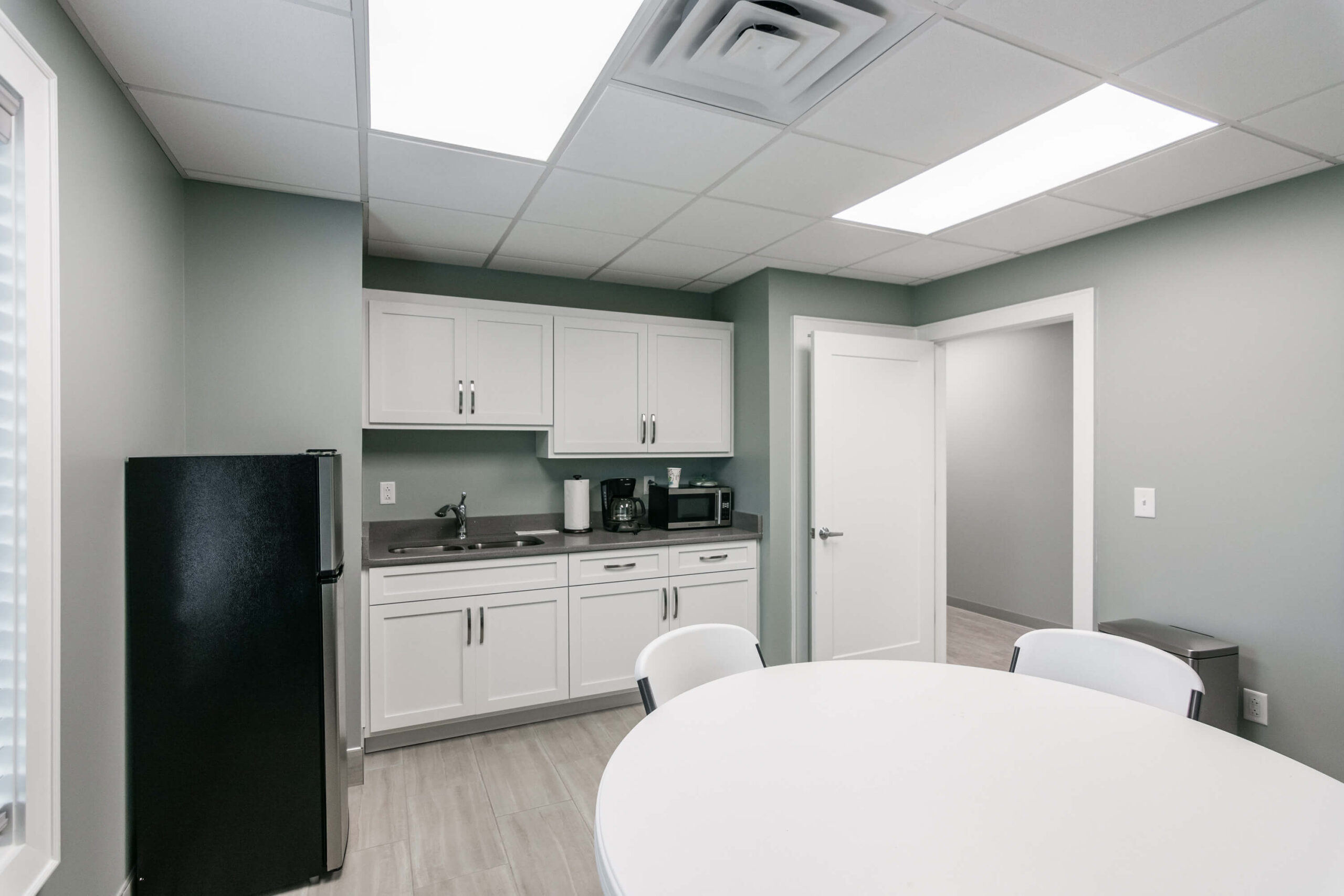 Small kitchenette with white cabinets, a black refrigerator, sink, microwave, and a round table with chairs. Green walls and tiled floor. Bright lighting from ceiling fixtures.