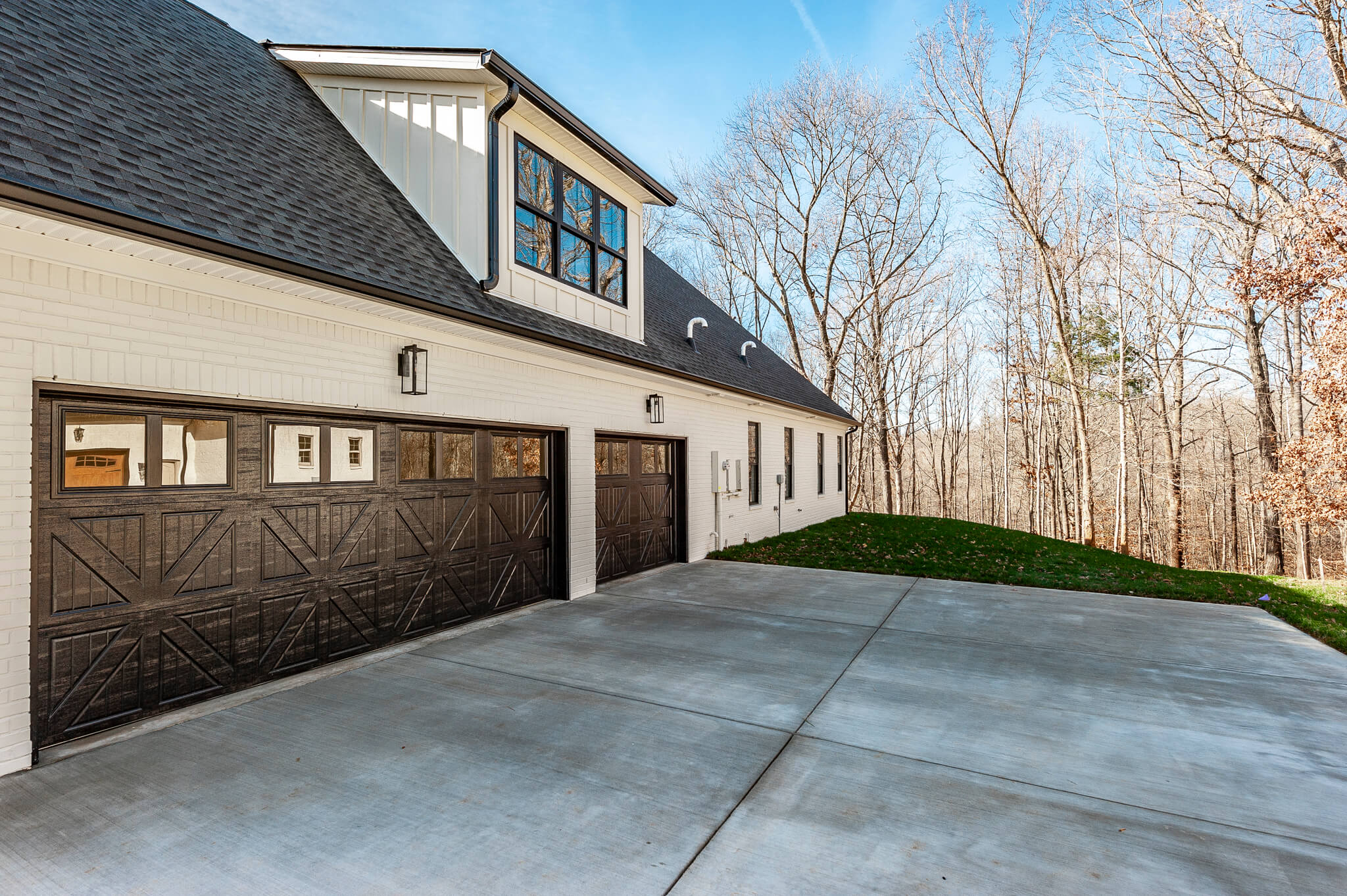 A two-story white house with three-car garage doors, set on a concrete driveway. Bare trees surround the property under a clear sky.